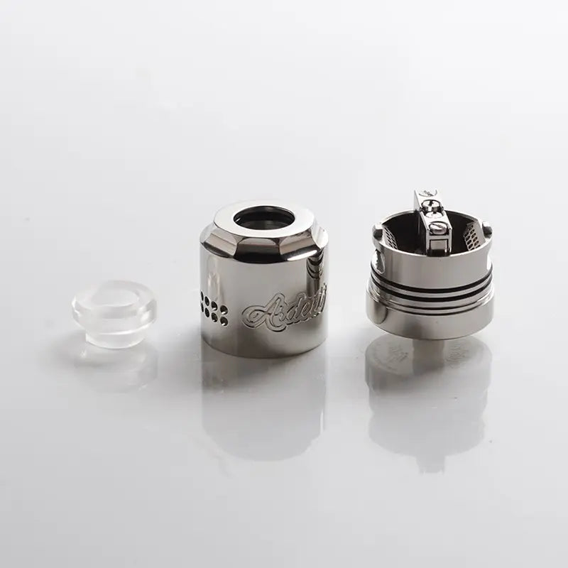 A silver metallic vape coil assembly shown disassembled on a reflective white surface, featuring a clear glass or acrylic mouthpiece piece on the left, with the main metal coil body and internal coil visible on the right.